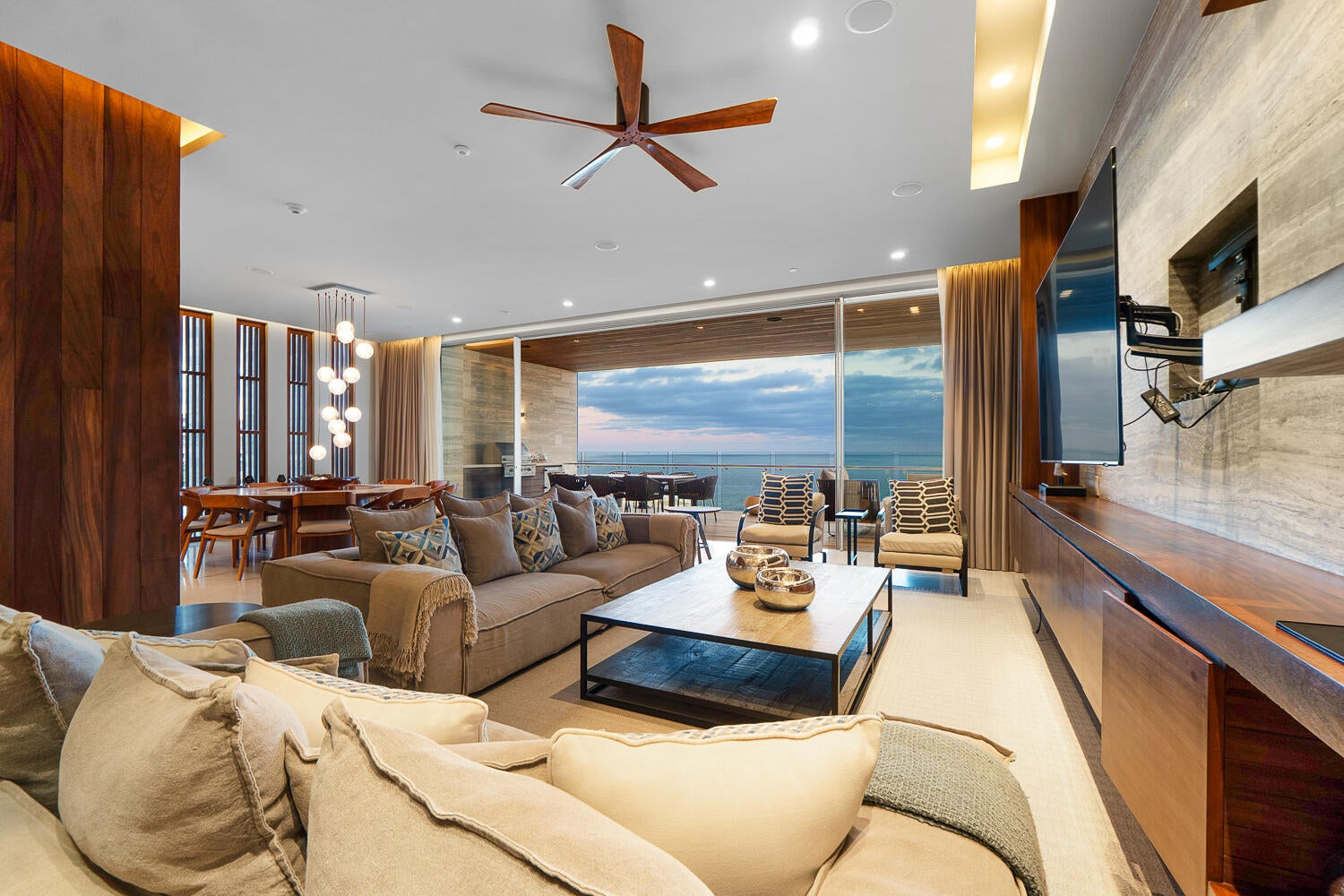 Open-plan living room flowing to ocean-view terrace at sunset, warm wood accents, designer furniture