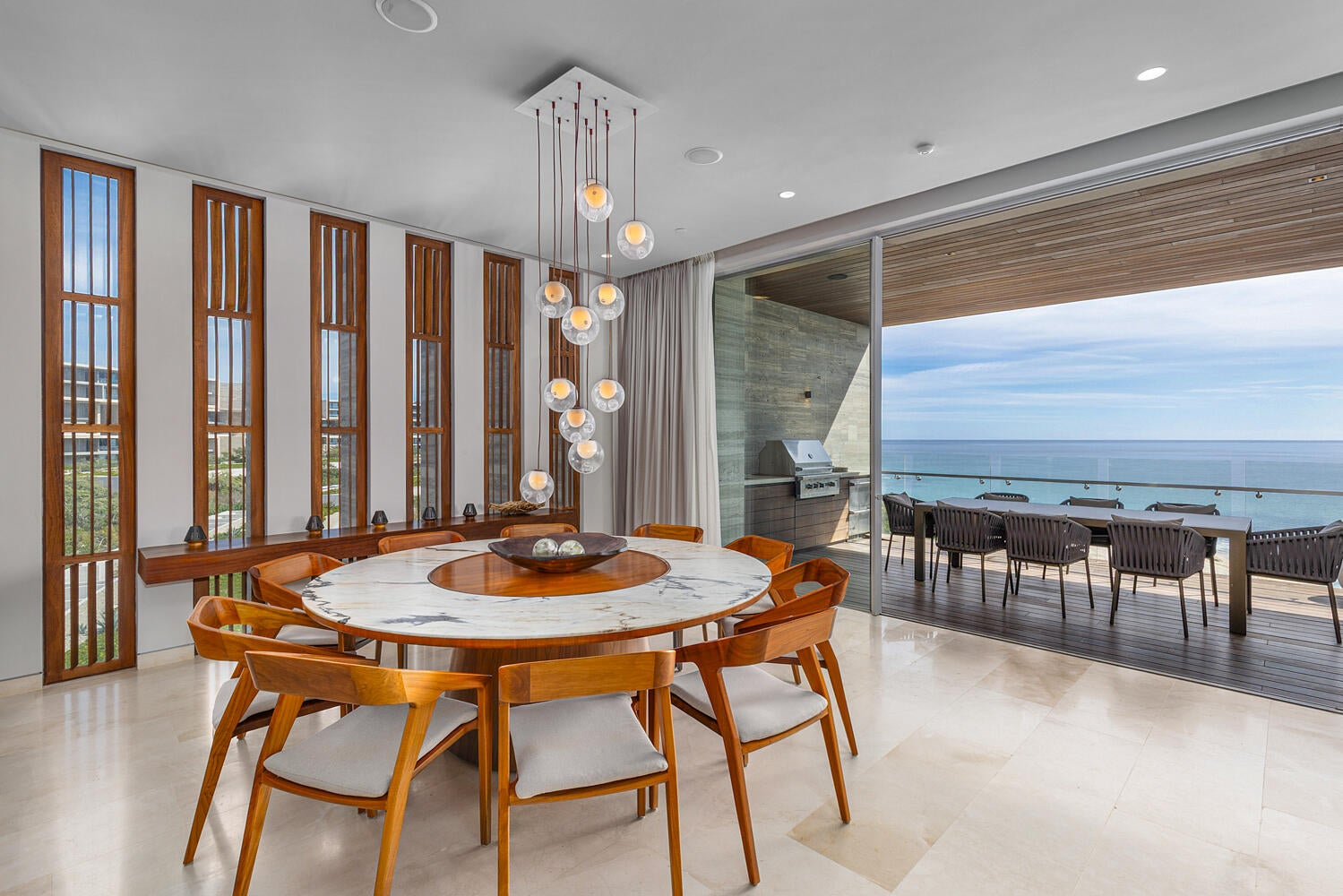 Dining area with round marble table, cascading glass pendant lights, floor-to-ceiling wood-slatted windows, ocean panorama