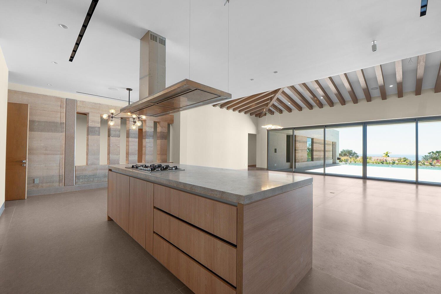 Modern minimalist kitchen with massive concrete-topped island, warm wood cabinetry, exposed geometric wood beam ceiling, glass walls opening to pool area