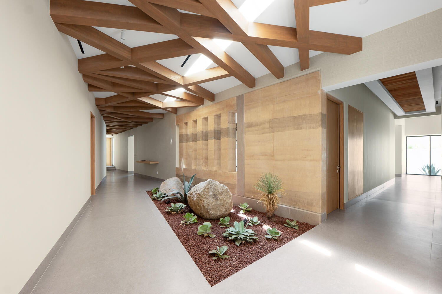 Interior courtyard with sunken desert garden — rocks, succulents, agave plants surrounded by warm wood walls and geometric exposed beams casting dramatic shadows