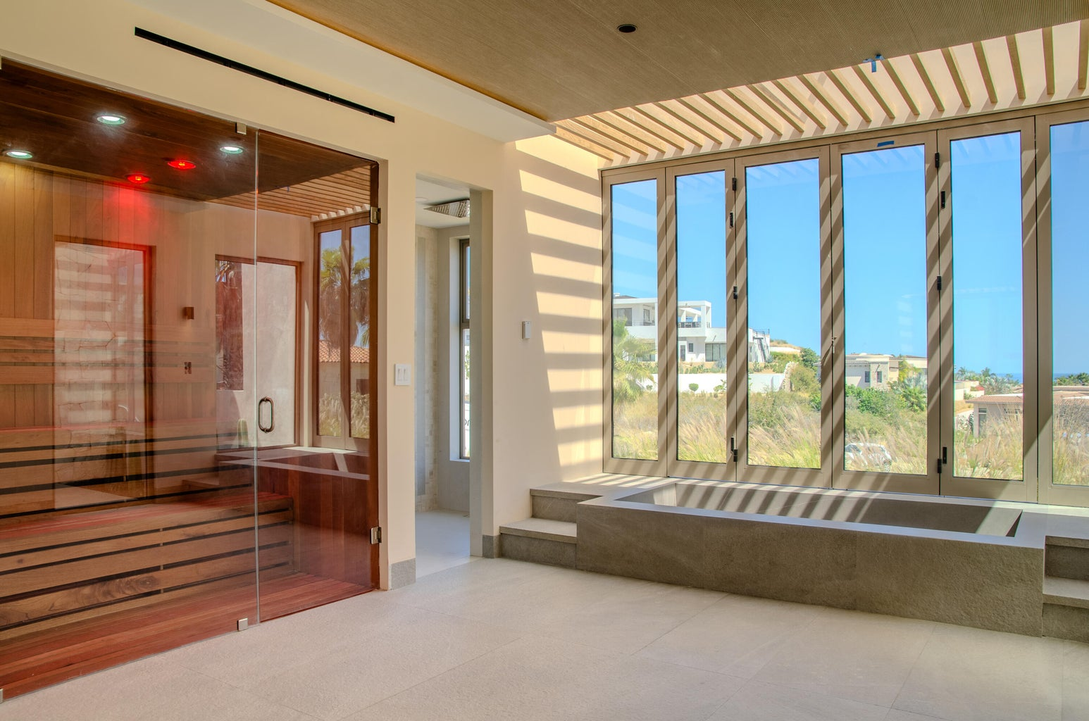 Wellness room with glass-enclosed red light therapy sauna, cedar interior with red glow, adjacent soaking tub, louvered windows casting striped shadow patterns